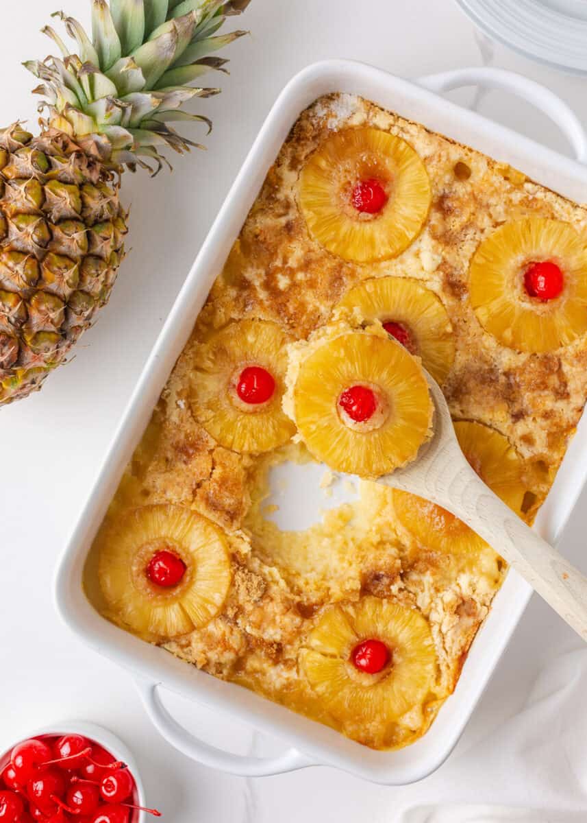 Overhead view of baked pineapple dump cake in a white casserole dish with pineapple rings, cherries, and a serving spoon.