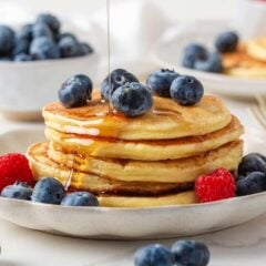 Stack of fluffy cottage cheese pancakes topped with fresh blueberries and maple syrup, served with raspberries on a ceramic plate.