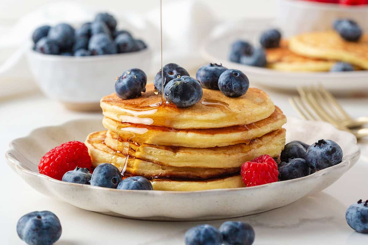 Stack of fluffy cottage cheese pancakes topped with fresh blueberries and maple syrup, served with raspberries on a ceramic plate.