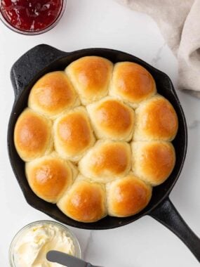 Overhead view of soft, golden dinner rolls baked in a cast iron skillet with butter and jam.