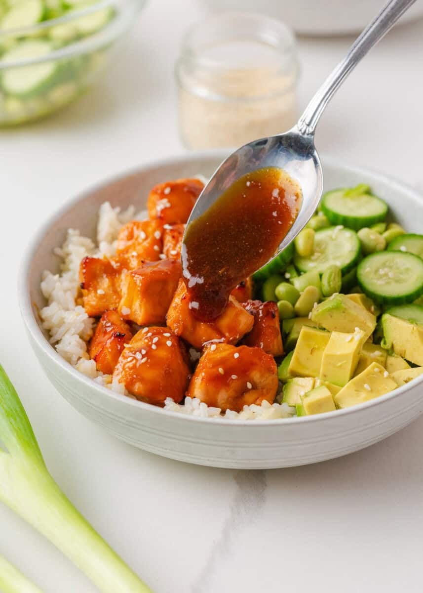 Teriyaki glaze being spooned over broiled salmon bites in a bowl with white rice, diced avocado, cucumber slices, and edamame.