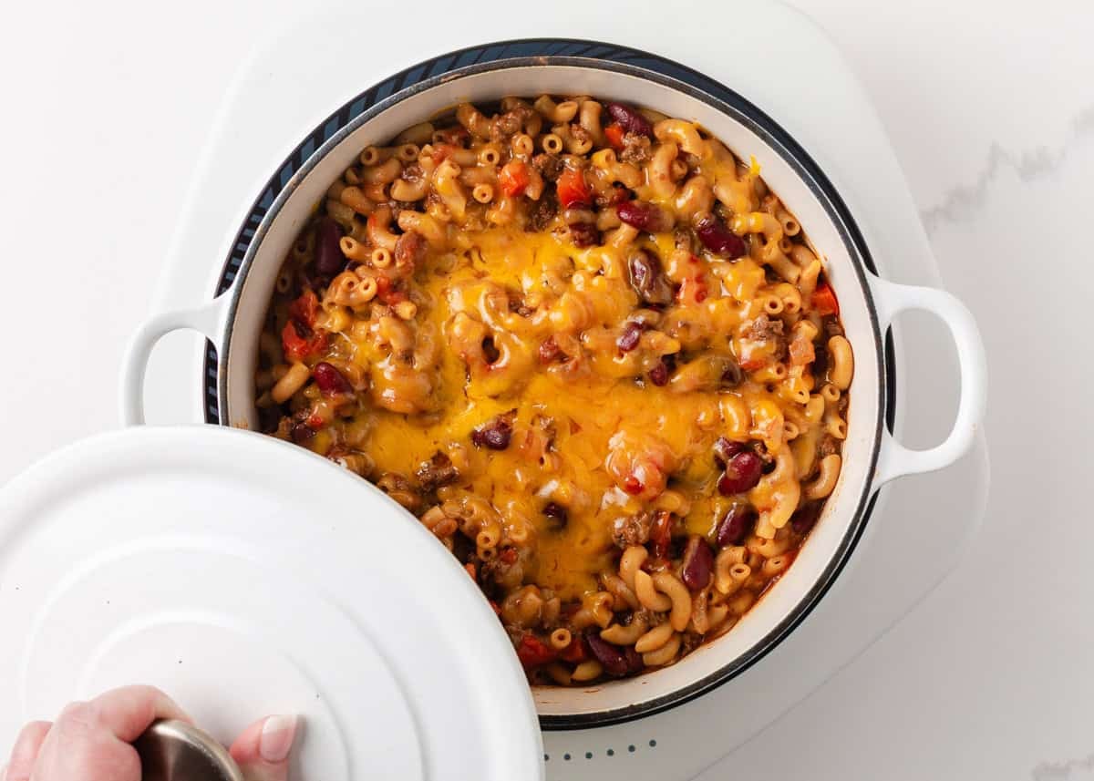 Finished cheesy chili mac revealed in a white Dutch oven with melted cheddar cheese on top.