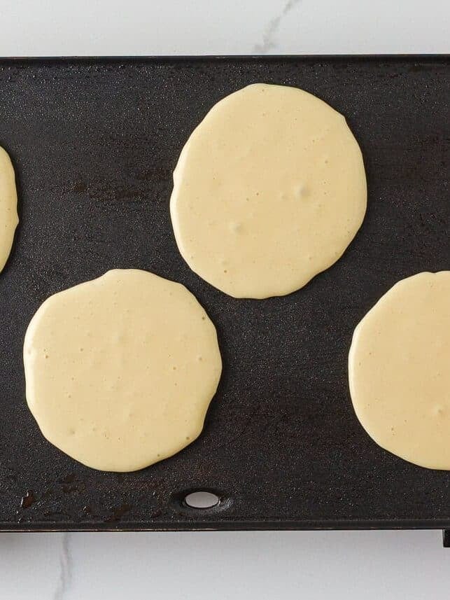 Four cottage cheese pancakes cooking on a buttered griddle until bubbles form on the surface.