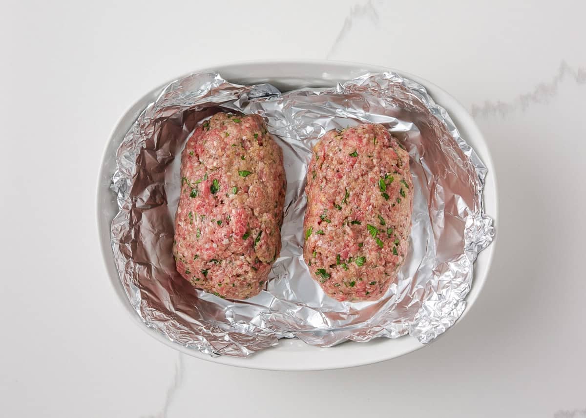 Two uncooked mini meatloaves shaped and placed in a foil-lined baking dish.