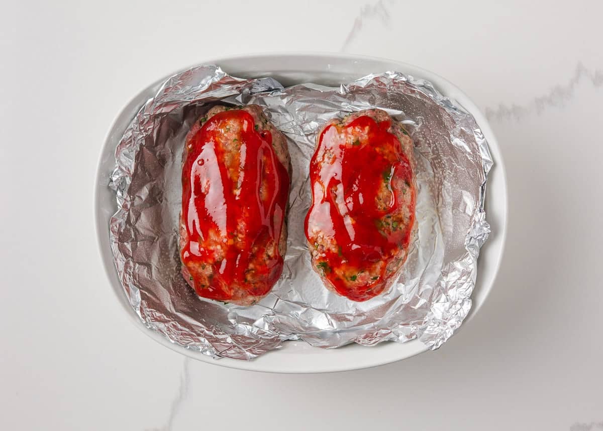 Two mini meatloaves brushed with ketchup glaze and ready to bake in a foil-lined dish.