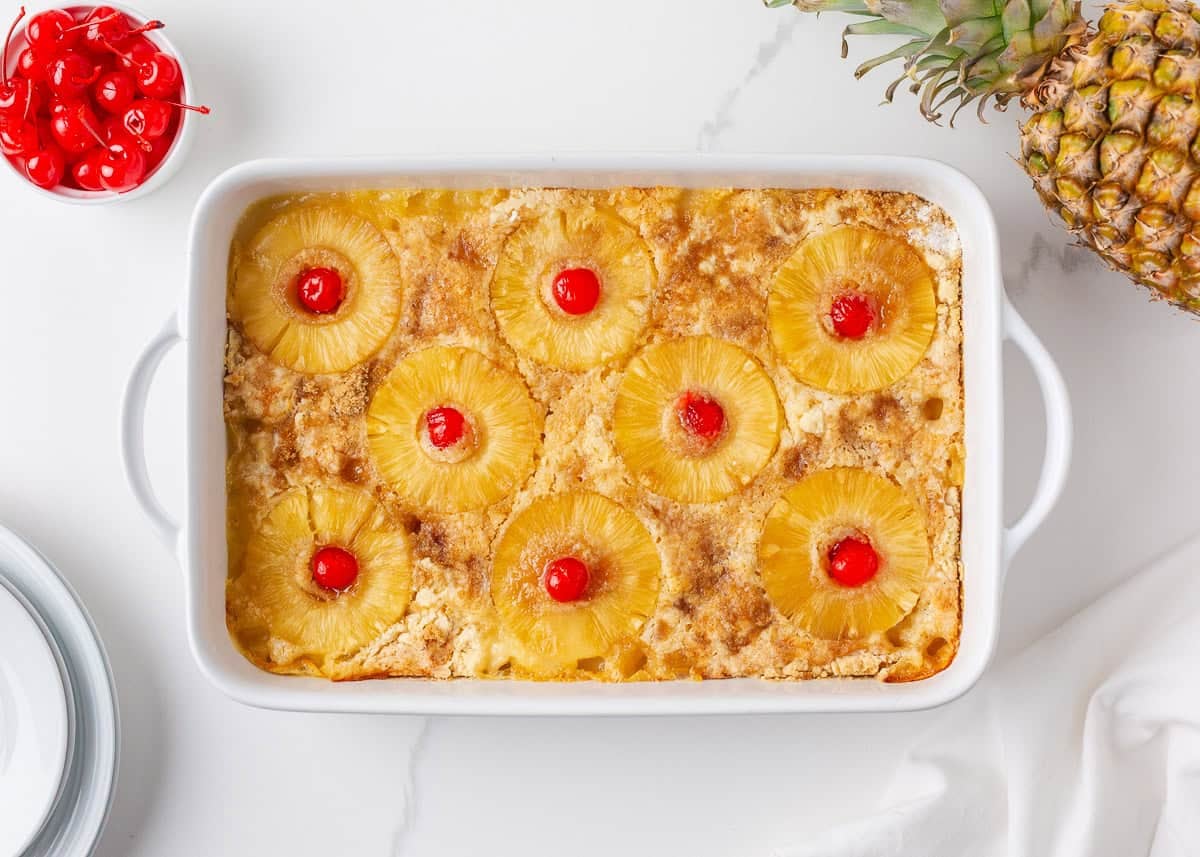 Baked pineapple dump cake with golden topping, pineapple rings, and cherries in a white baking dish.