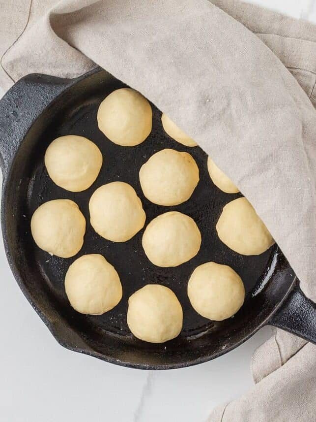 Twelve shaped dinner roll dough balls rising in a cast iron skillet under a kitchen towel.