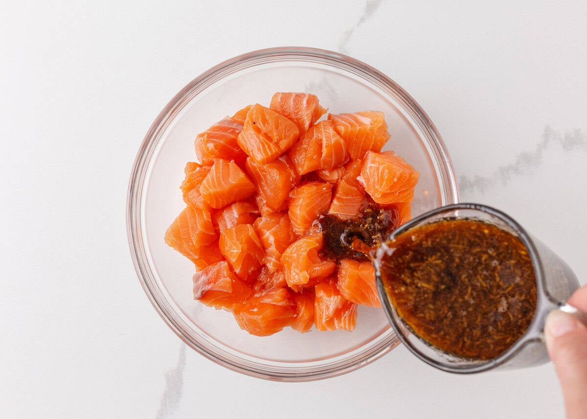 Homemade teriyaki marinade being poured over cubed salmon in a glass bowl.