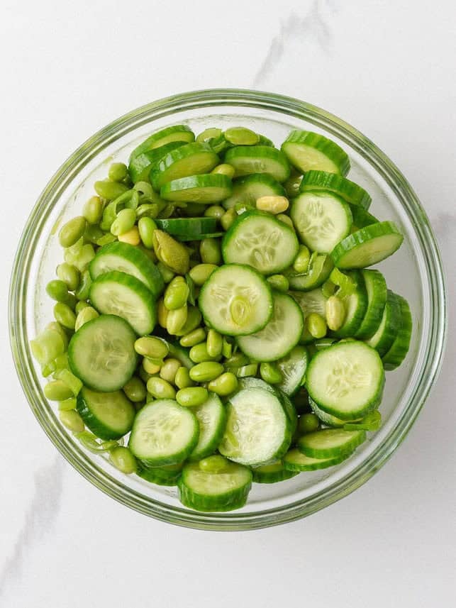 Cucumber and edamame salad with sliced green onions in a glass mixing bowl.
