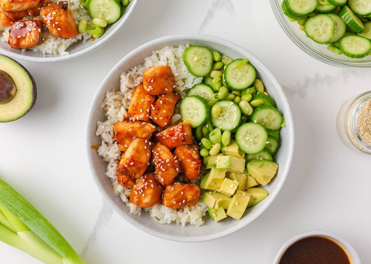 Teriyaki salmon rice bowl with cucumber, edamame, avocado, green onions and sesame seeds.