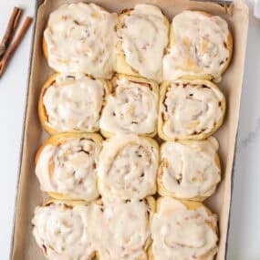 Apple cinnamon rolls topped with maple icing on a parchment-lined baking sheet with green apples and cinnamon sticks.