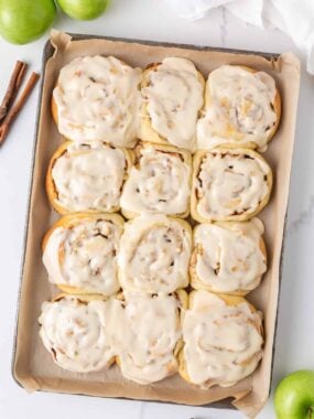 Apple cinnamon rolls topped with maple icing on a parchment-lined baking sheet with green apples and cinnamon sticks.