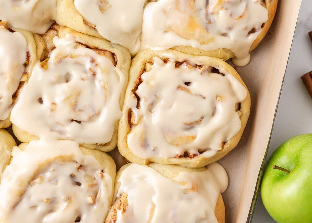Close-up of soft apple cinnamon rolls covered with creamy maple icing beside a green apple.