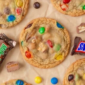 Soft baked candy cookies loaded with colorful M&M&rsquo;s and chopped candy bar pieces on parchment paper.