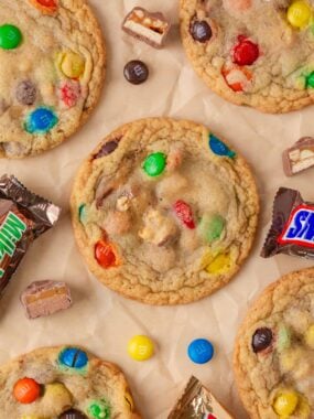 Soft baked candy cookies loaded with colorful M&M&rsquo;s and chopped candy bar pieces on parchment paper.