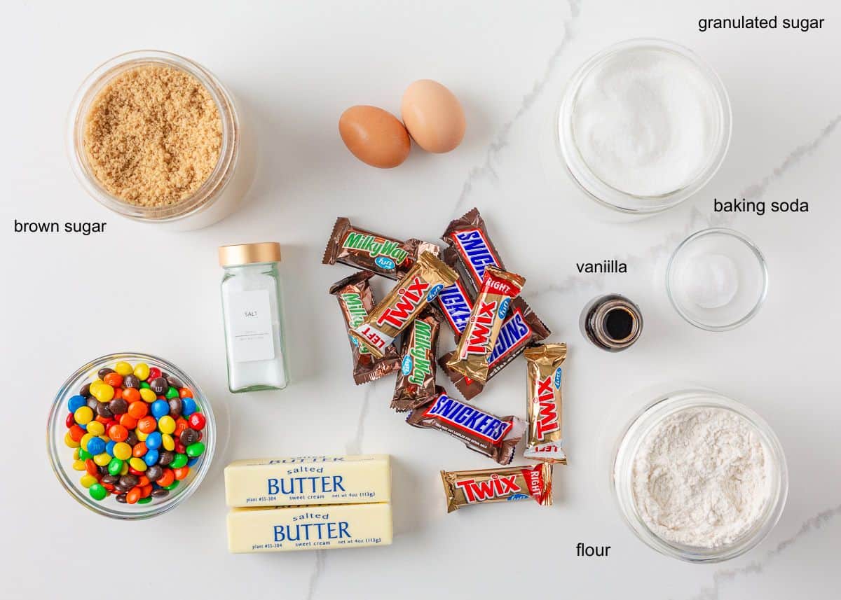 Ingredients for candy cookies including M&M’s, chopped candy bars, butter, sugars, eggs, vanilla, flour, baking soda and salt.