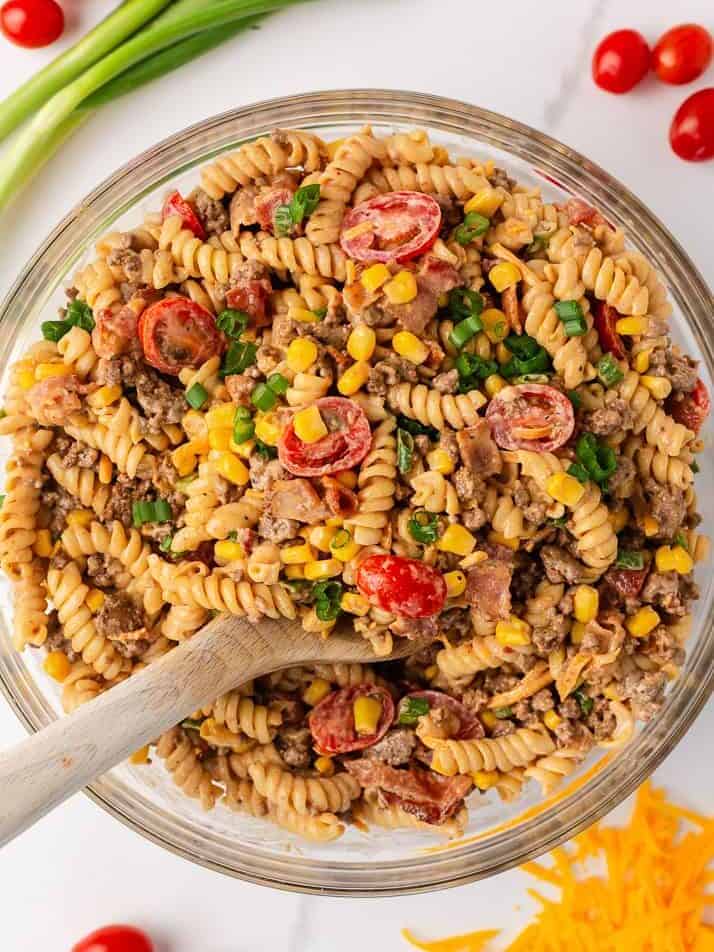 Overhead view of a large glass bowl of cowboy pasta salad with rotini, ground beef, corn, cherry tomatoes, bacon, cheddar cheese, and green onions.