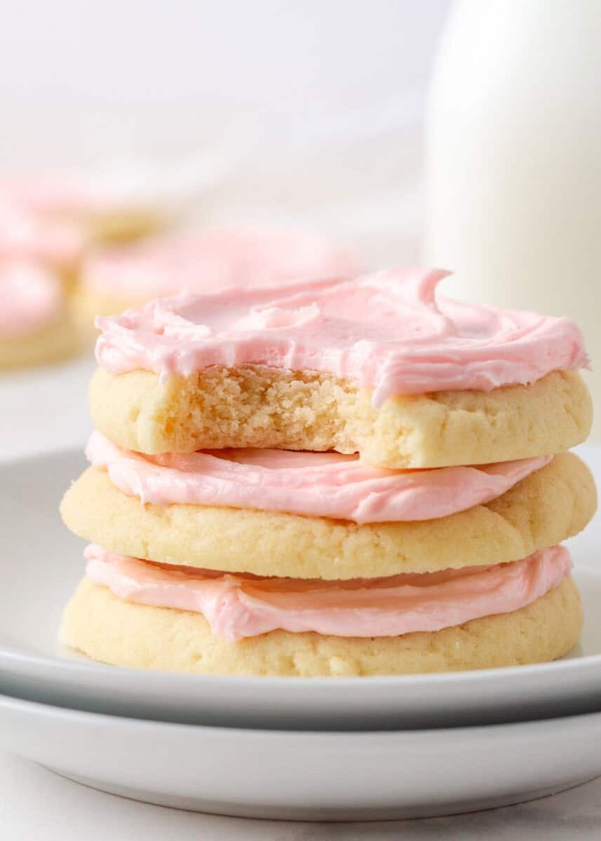 Stack of soft Crumbl sugar cookies topped with pink almond frosting, with a bite taken from the top cookie.