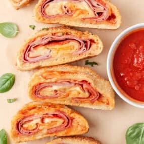 Sliced homemade stromboli filled with melted cheese, ham, salami and pepperoni beside marinara sauce.