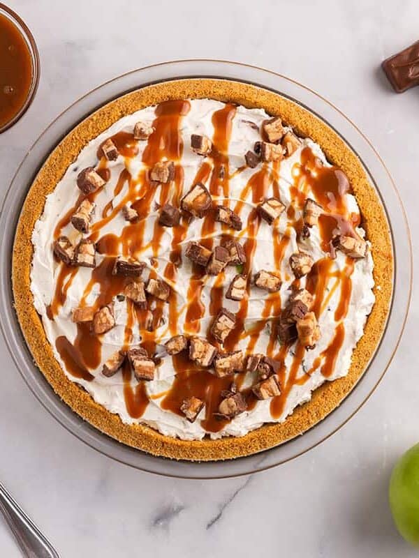 Snickers pie topped with chopped Snickers and caramel sauce in a graham cracker crust, surrounded by green apples and candy bars.