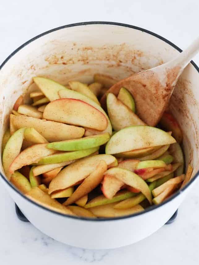 Apple slices tossed with cinnamon sugar in a pot with a wooden spoon.