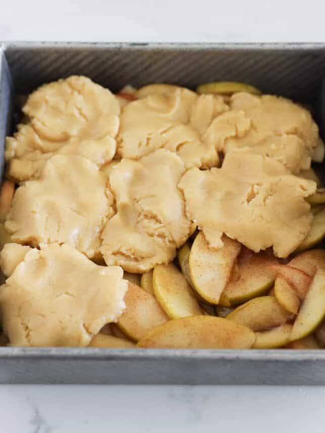 Cobbler dough flattened over cinnamon apple filling in a square baking pan.