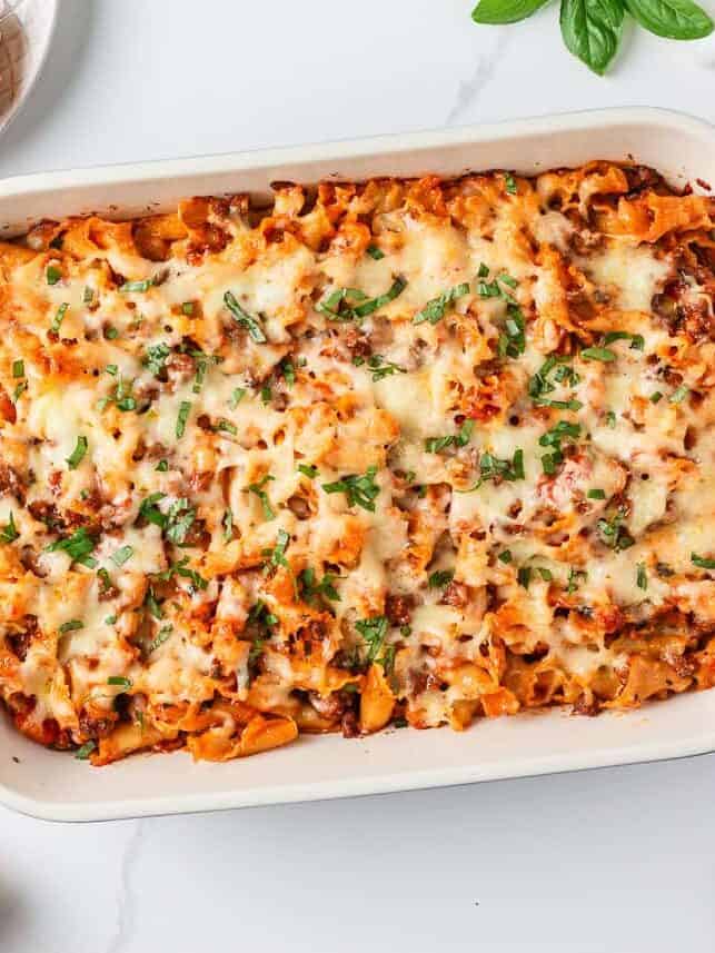 Overhead view of an easy lasagna casserole in a white baking dish with golden melted cheese, surrounded by fresh basil and Parmesan.