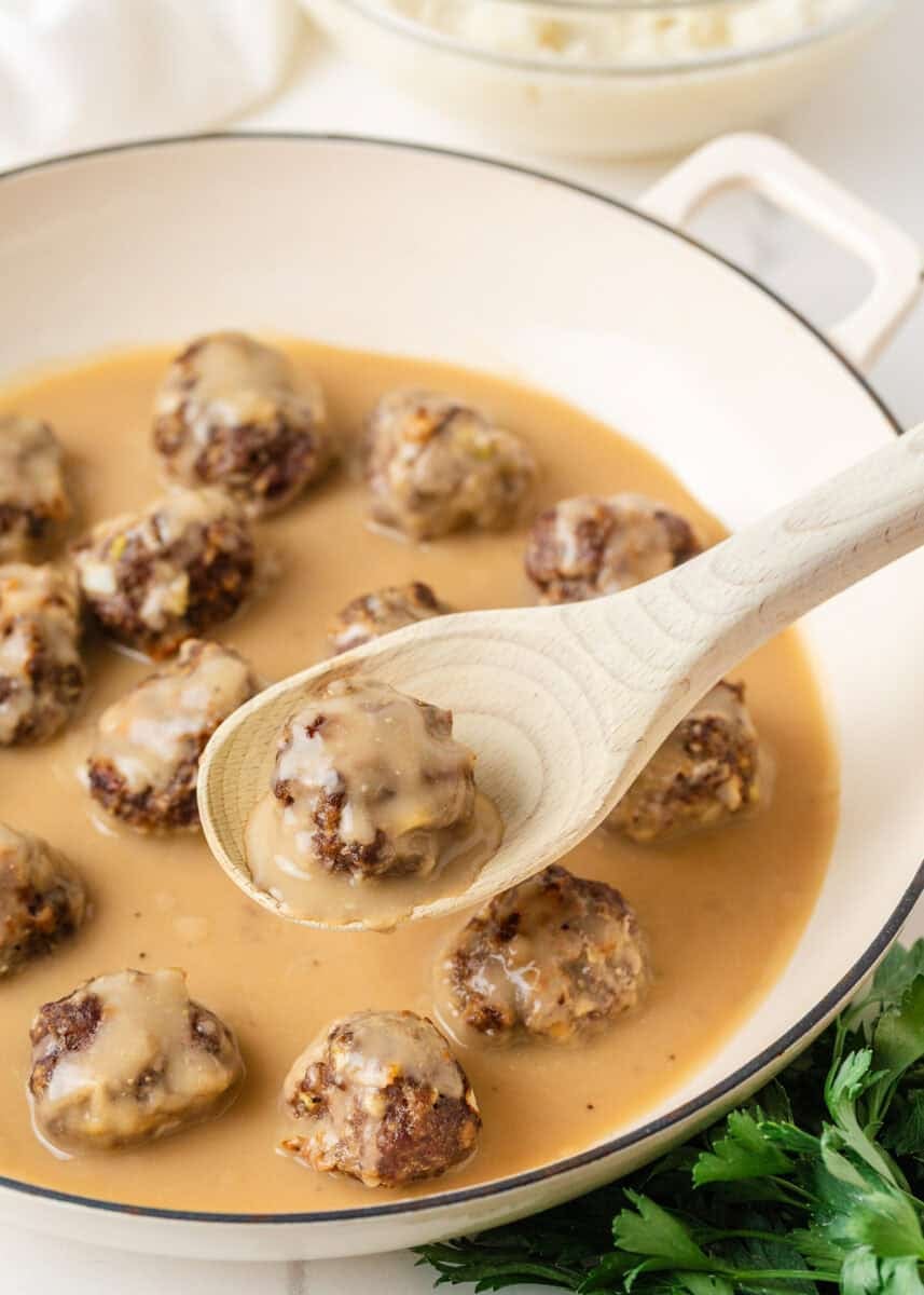 Homemade meatballs simmering in brown gravy with one meatball lifted on a wooden spoon.