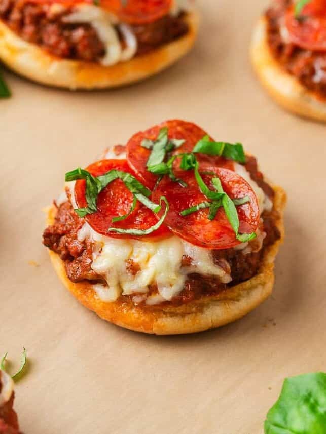 Close-up of mini open-faced pizza burgers with melted cheese, pepperoni slices, and fresh basil on toasted buns.