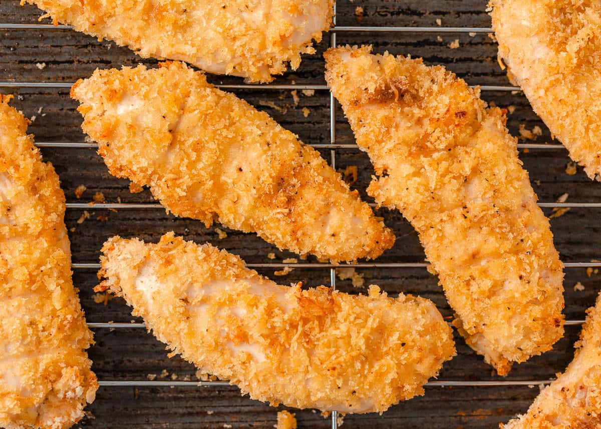 These baked chicken tenders are perfectly crispy on the outside and tender on the inside. A family-favorite recipe that's simple, delicious and ready in just 30 minutes! Close-up of golden crispy baked chicken tenders on a wire cooling rack.