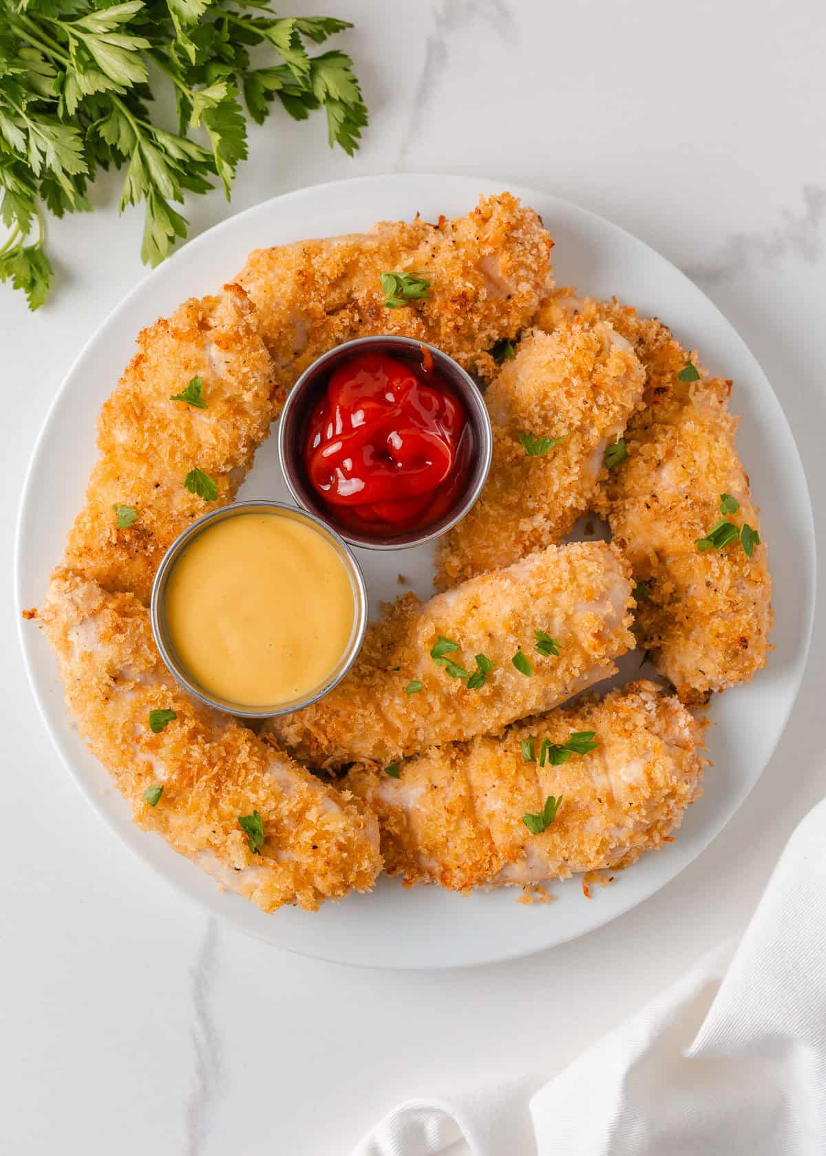 These baked chicken tenders are perfectly crispy on the outside and tender on the inside. A family-favorite recipe that's simple, delicious and ready in just 30 minutes! Crispy baked chicken tenders served with ketchup and dipping sauce on a white plate.