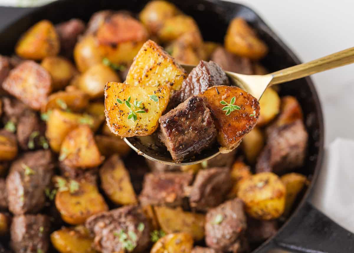 Spoonful of garlic butter steak bites and golden potatoes held over a cast iron skillet.