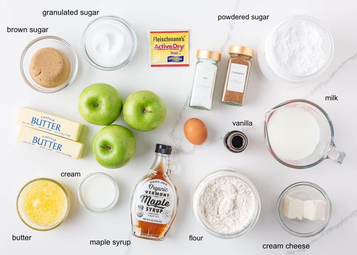 Apple cinnamon roll ingredients including flour, yeast, brown and granulated sugar, cinnamon, salt, green apples, butter, egg, milk, cream cheese, powdered sugar, vanilla and maple syrup.