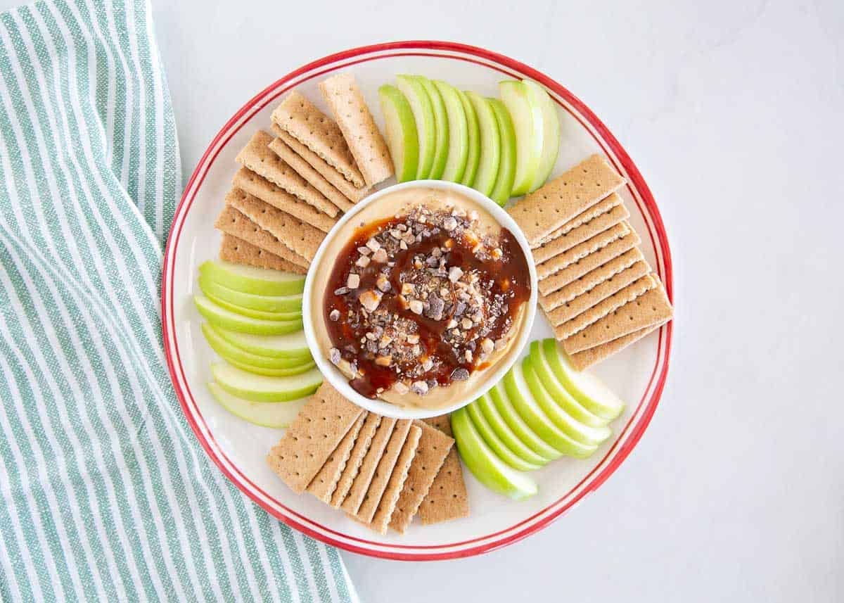 Caramel apple cheesecake dip on a platter surrounded by green apple slices and graham crackers.