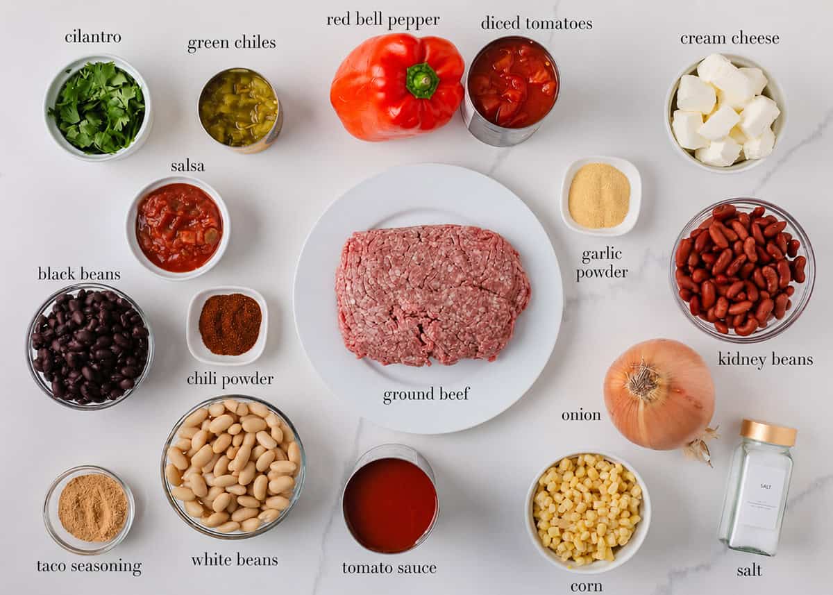Creamy taco soup ingredients arranged on a white countertop, including ground beef, three types of beans, corn, tomatoes, peppers, onion, cream cheese and seasonings.