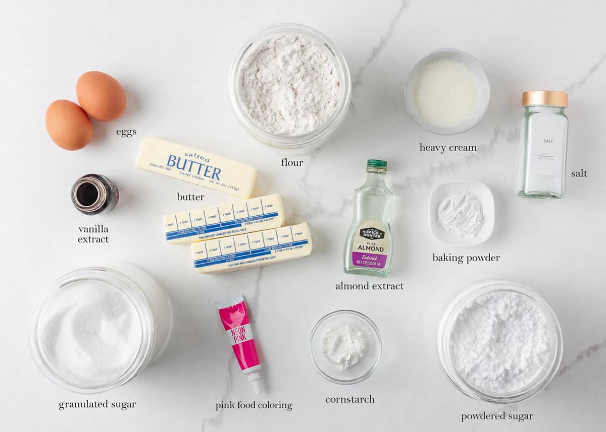 Overhead view of labeled ingredients for copycat Crumbl sugar cookies and pink almond frosting, including butter, eggs, flour, sugar, extracts and pink food coloring.