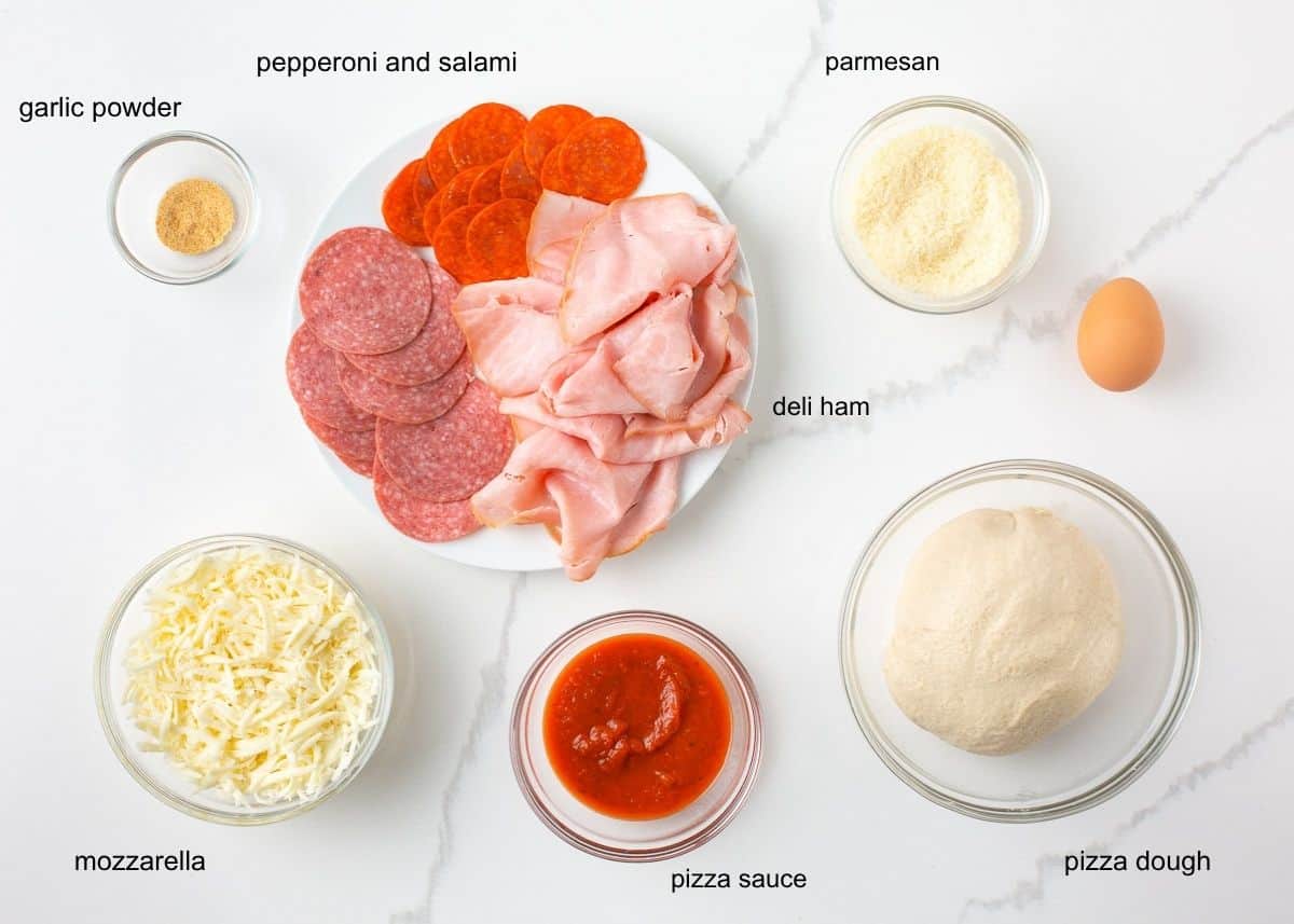 Ingredients for homemade stromboli including pizza dough, mozzarella, pizza sauce, ham, salami, pepperoni, Parmesan, egg and garlic powder.