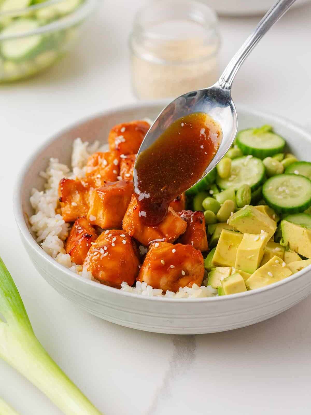 Teriyaki glaze being spooned over broiled salmon bites in a bowl with white rice, diced avocado, cucumber slices, and edamame.