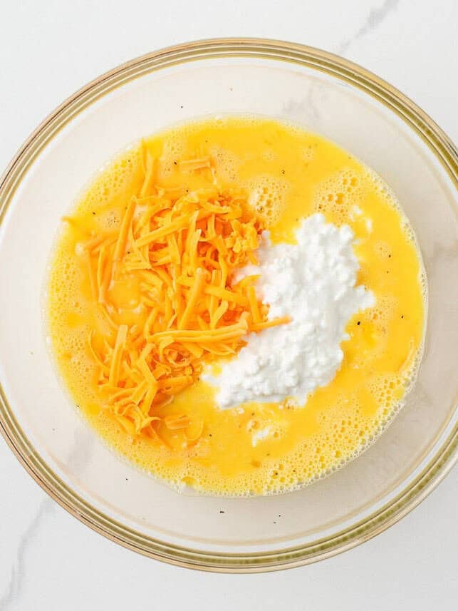 Overhead view of a large glass mixing bowl with whisked eggs and milk, topped with shredded cheddar cheese and a scoop of small curd cottage cheese, before stirring to combine.
