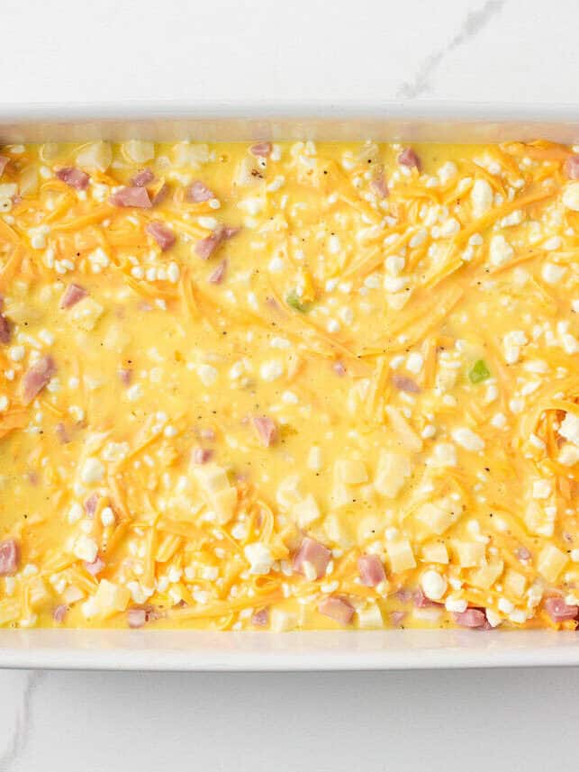 Overhead view of a white baking dish filled with the unbaked egg, cheese, ham, and potato mixture, ready to go into the oven.