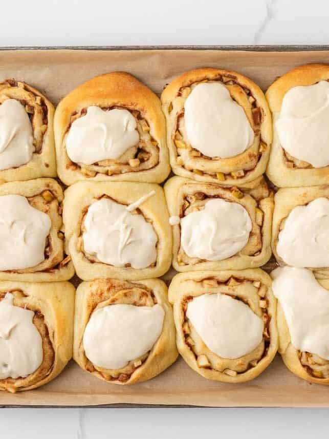 Freshly baked apple cinnamon rolls topped with dollops of maple icing on a baking sheet.