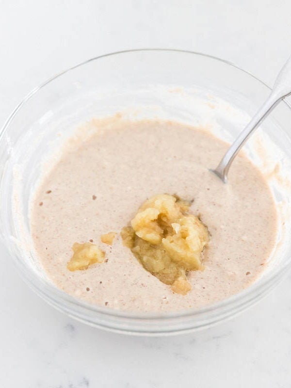 Grated apple and applesauce added to cinnamon pancake batter in a glass mixing bowl.