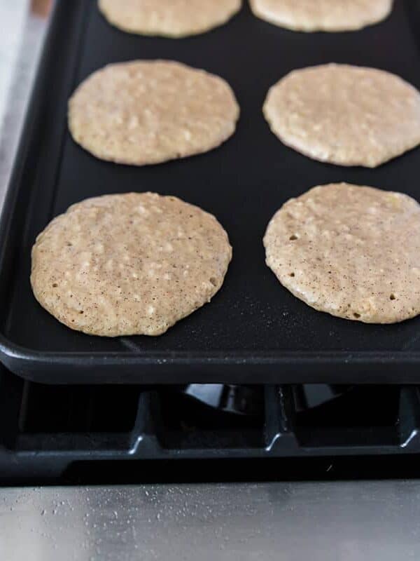 Apple pancakes cooking on a stovetop griddle until bubbles form on the surface.