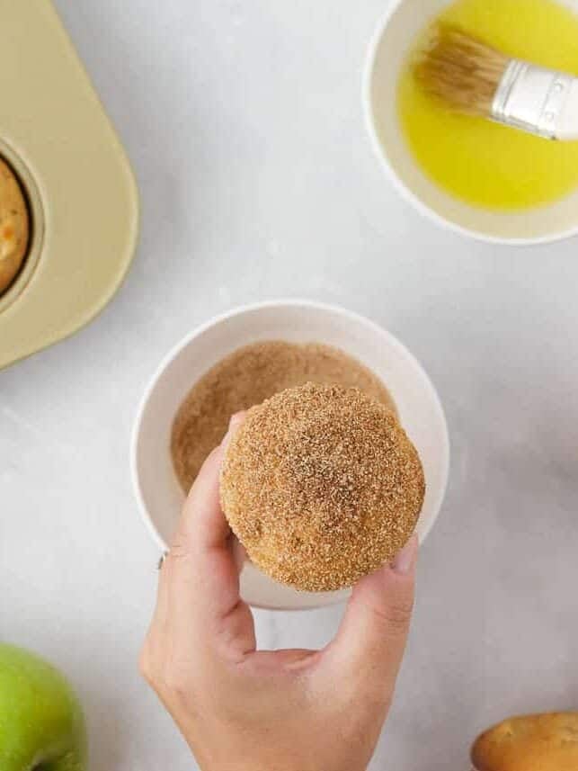 Hand holding a freshly baked apple muffin coated in cinnamon sugar.