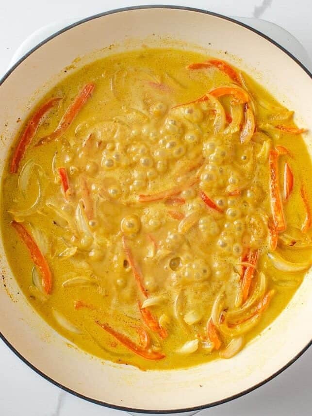 Coconut curry sauce simmering with sliced red bell peppers and onions in a Dutch oven.