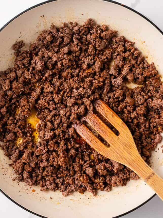 Seasoned ground beef browning in a Dutch oven, broken into crumbles with a wooden spatula.