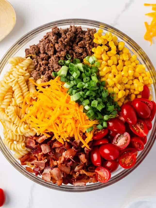 Cowboy pasta salad ingredients arranged in sections in a large glass bowl before tossing.
