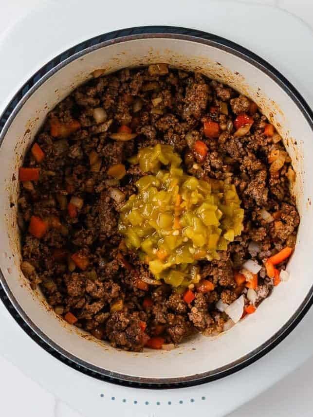 Ground beef cooked with onion and red bell pepper, topped with diced green chiles in a Dutch oven.