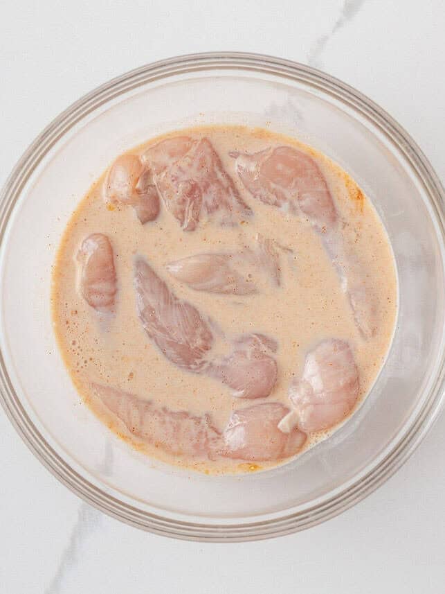 Chicken tenderloins marinating in seasoned buttermilk in a glass bowl.