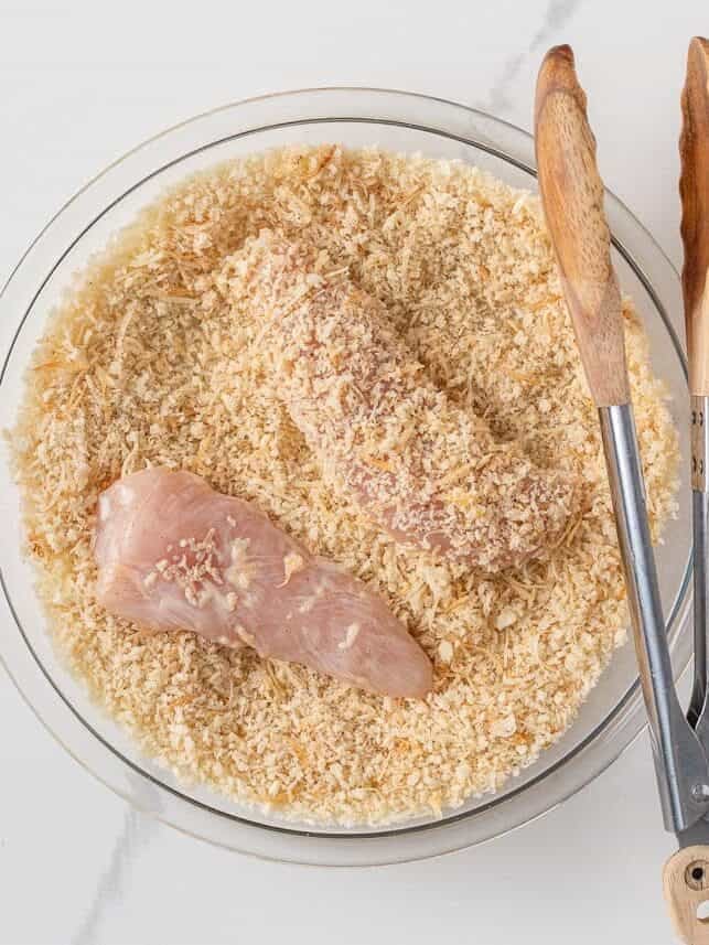 Chicken tenderloins being coated in seasoned panko breadcrumbs with kitchen tongs.